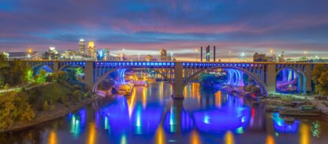 Minneapolis skyline and I-35W bridge lit up at night, reflecting over the Mississippi River.
